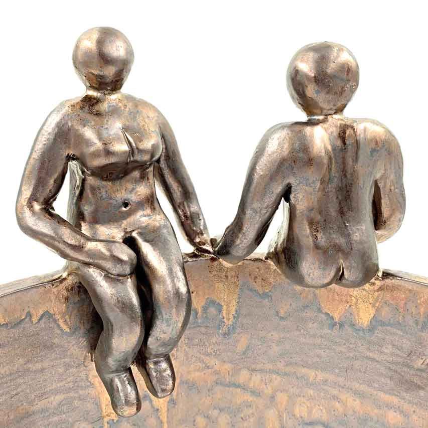 Twee kleine, bronskleurige figuurtjes zitten op een gebogen richel, de ene kijkt naar voren en de andere is afgewend.