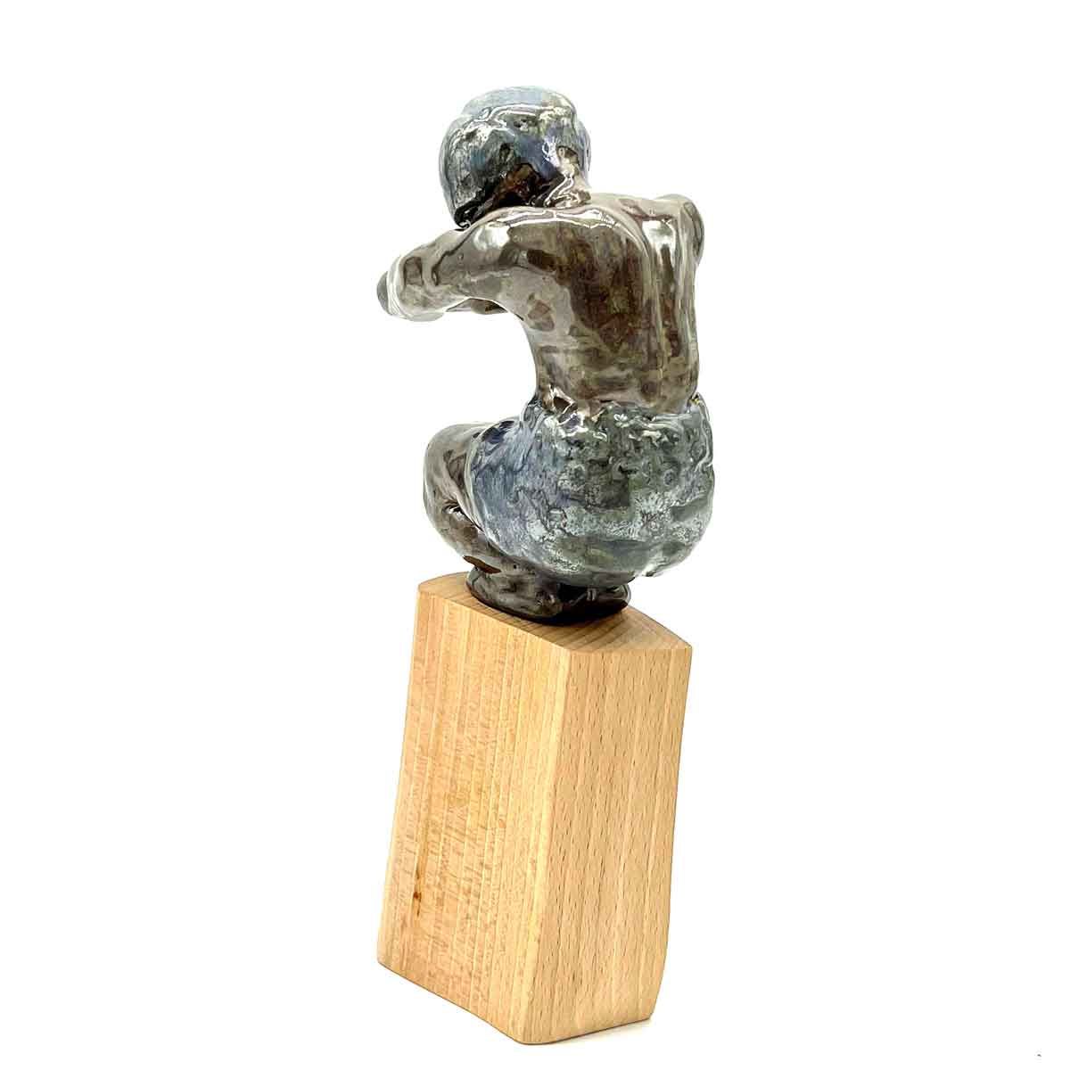 Klein grijs metalen beeldje op een houten blokje, van achteren gezien in een opgerolde zittende houding.