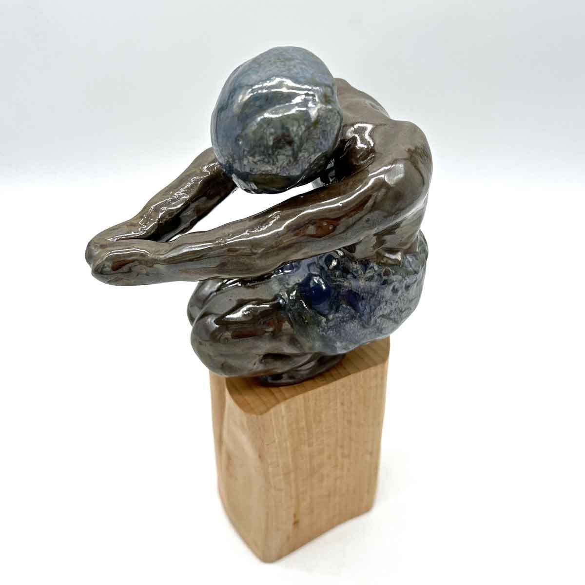 Abstracte blauwgrijze keramische figuur, gehurkt met uitgestrekte armen, op een houten voetstuk.