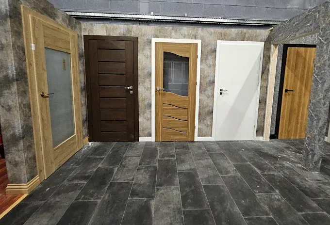 Five interior doors in different styles displayed in a showroom with tile flooring and textured walls.
