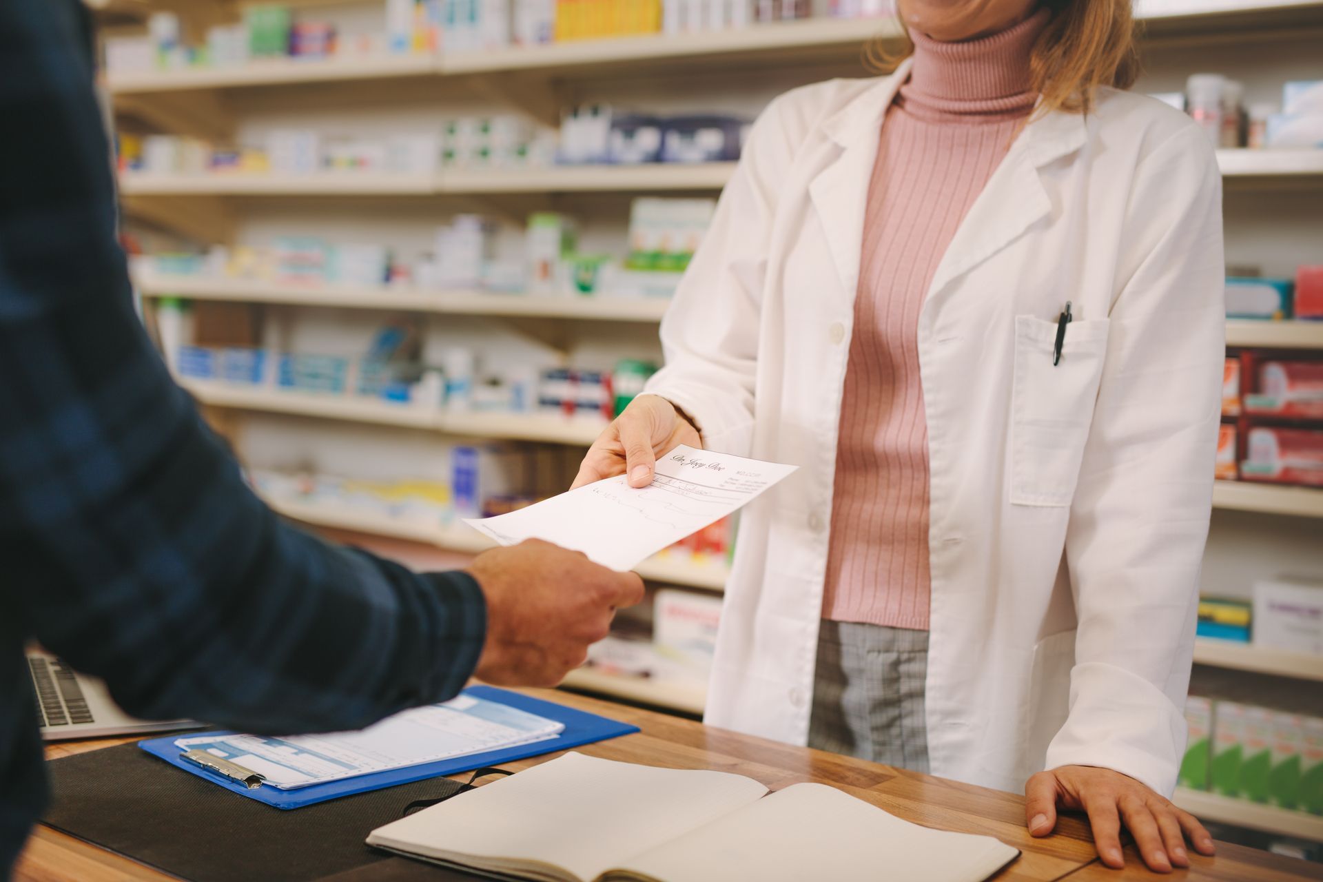 Ein Apotheker übergibt einem Kunden am Apothekenschalter ein Rezept; im Hintergrund sind Regale mit Medikamenten zu sehen.