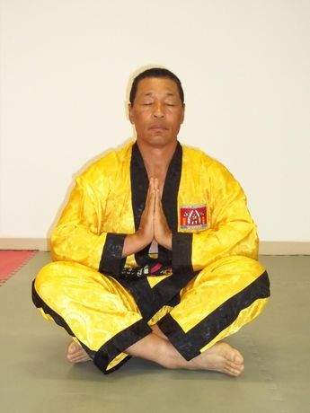 A man in a yellow and black karate uniform sits in a lotus position with his eyes closed