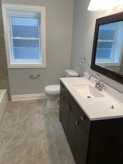 Bathroom with gray walls, dark vanity, white sink, toilet, mirror, and window with blinds.