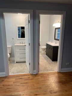 Hallway view into two bathrooms, one white and one dark vanity, with hardwood floor in foreground