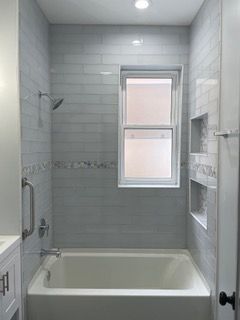 Accessible bathroom with white tiled shower-tub, grab bars, built-in shelves, and a window.