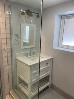 Small bathroom vanity beside a glass shower, with white cabinet and window