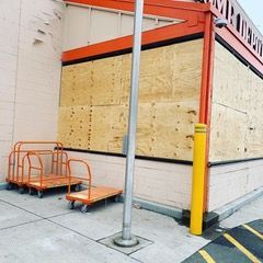 Boarded-up storefront with plywood windows, orange barricades, and a yellow safety post on the sidewalk