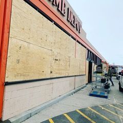 Boarded storefront with plywood over windows, sidewalk, and a blue cart outside.