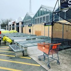 Outdoor market storefront with stacked metal trays, a shopping cart, and plywood-covered windows after damage