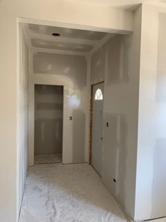 Renovated hallway with unfinished drywall, white walls, and a doorway leading to a lit room