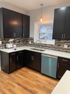 Modern kitchen with dark cabinets, white countertops, mosaic backsplash, and stainless dishwasher under a window.