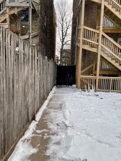 Snowy alleyway between wooden fences and apartment stairs, leading to a dark gate at the end