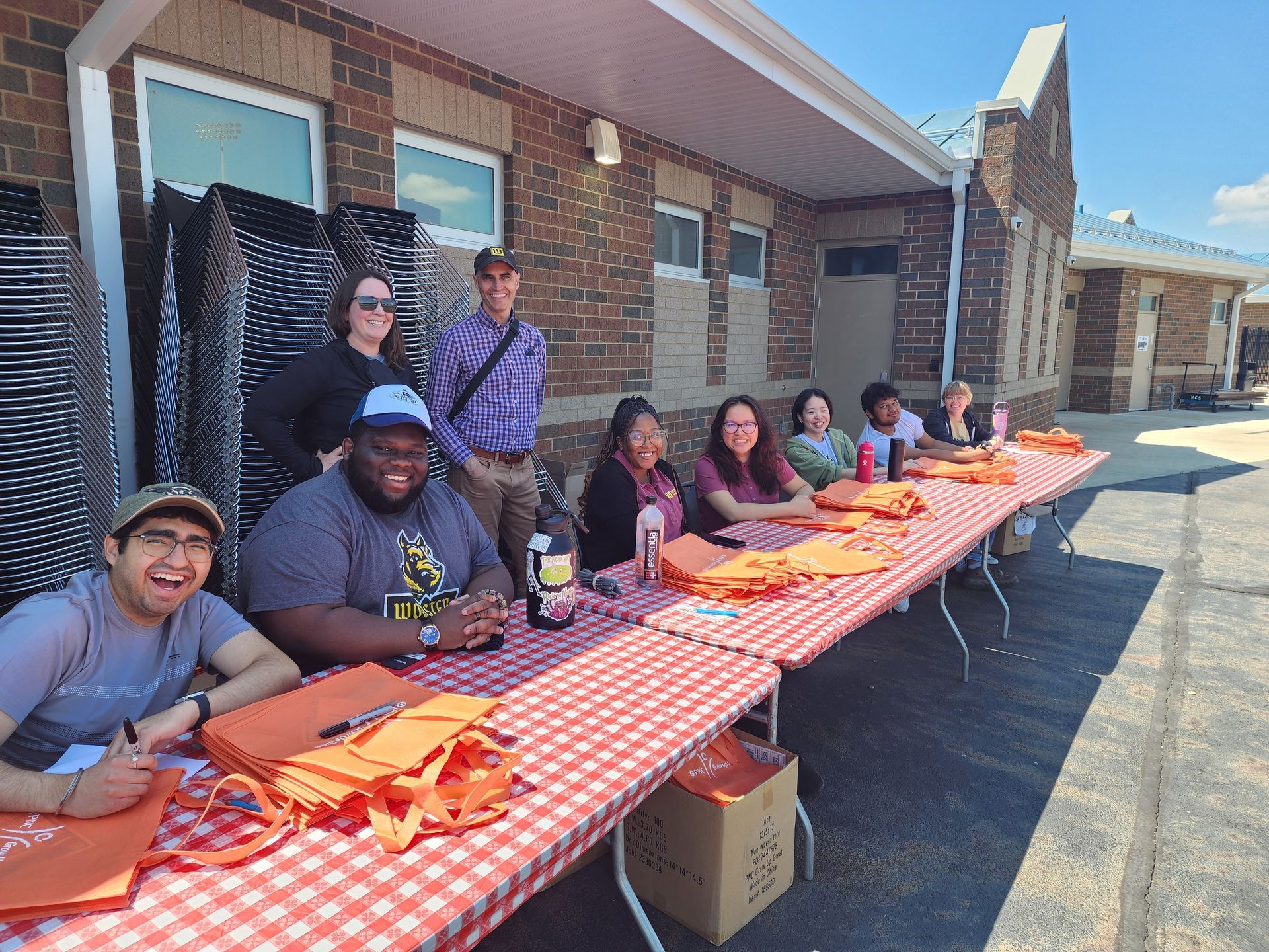 People seated at a table outside, smiling, with orange bags. A brick building is in the background.