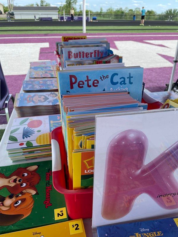 Books for sale on table at outdoor event, with purple sports field in background.