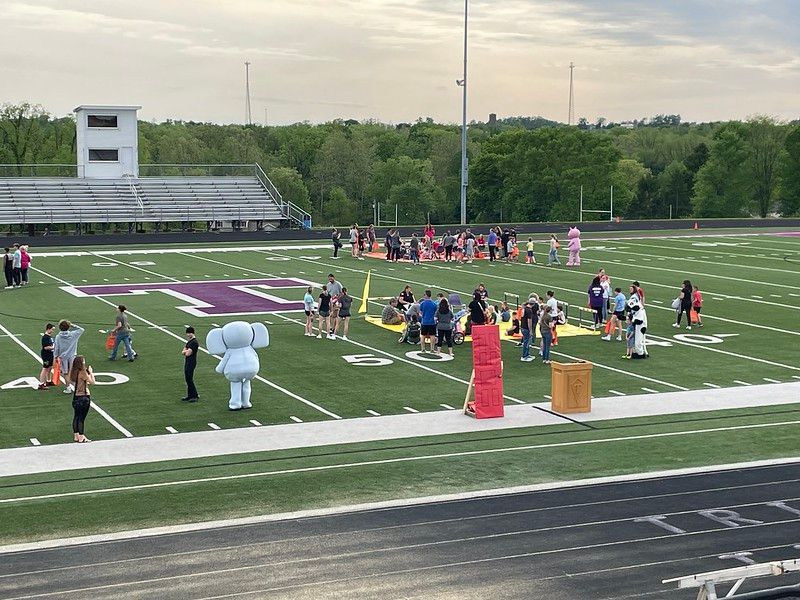 People gathered on a football field with mascots. A school is in the background.