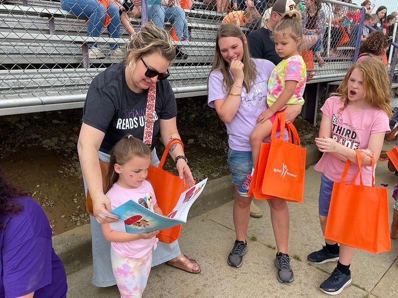 Woman reads a book to a young girl, others watch. They are holding orange bags near bleachers.