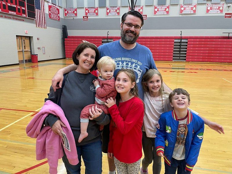 Family of six smiles together in a gymnasium. People have various expressions. Red and wood tones in the background.