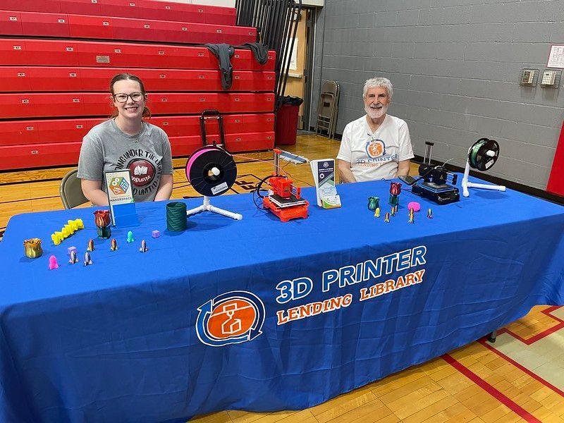Two people at a 3D printer display table, showcasing printed items.
