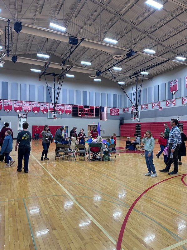 People gathered in a gymnasium with a basketball court. Red and white banners decorate the walls.