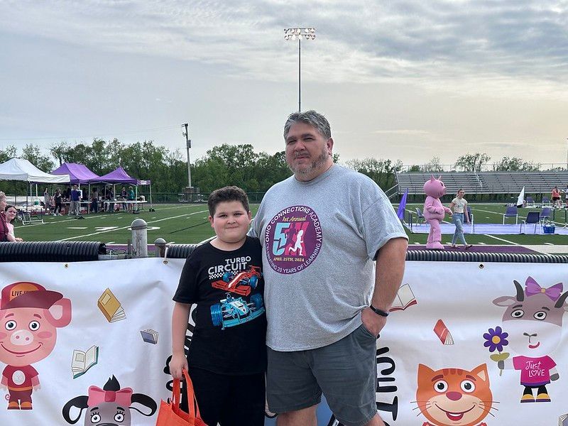 Father and son pose in front of a banner at an outdoor event. A pig and cat cartoon figures are visible.