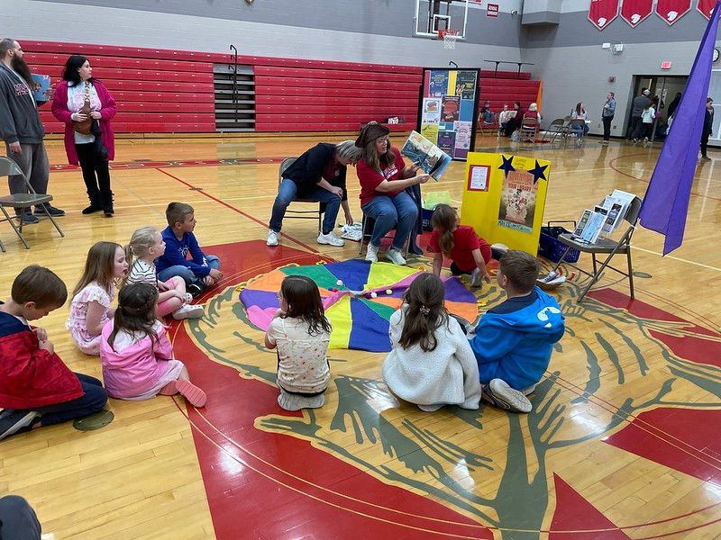 Children sit on gym floor listening to a woman read aloud. Colorful mat, books, and props are present.