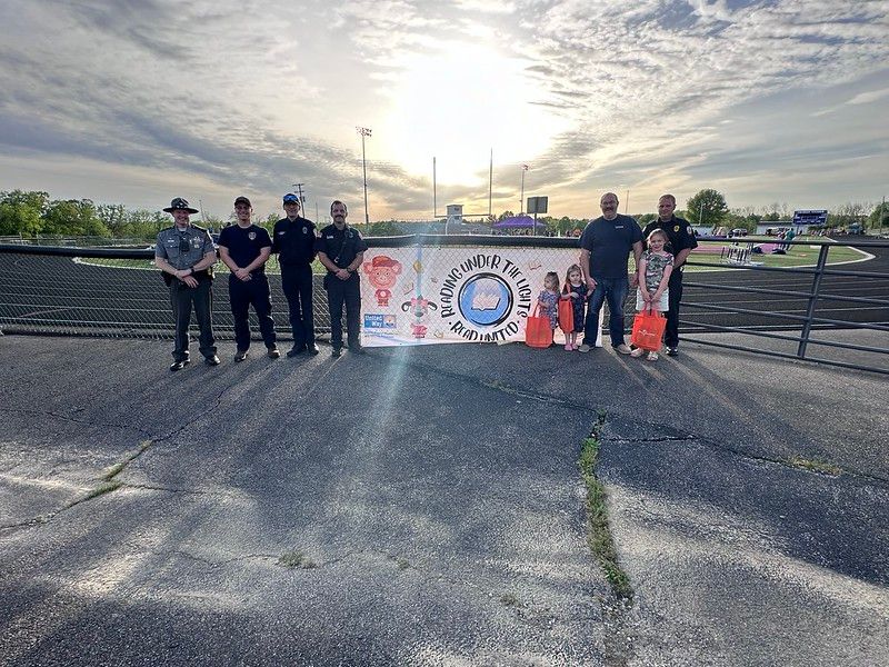 Group of people, including law enforcement and children, hold banner on a track with sun in the background.