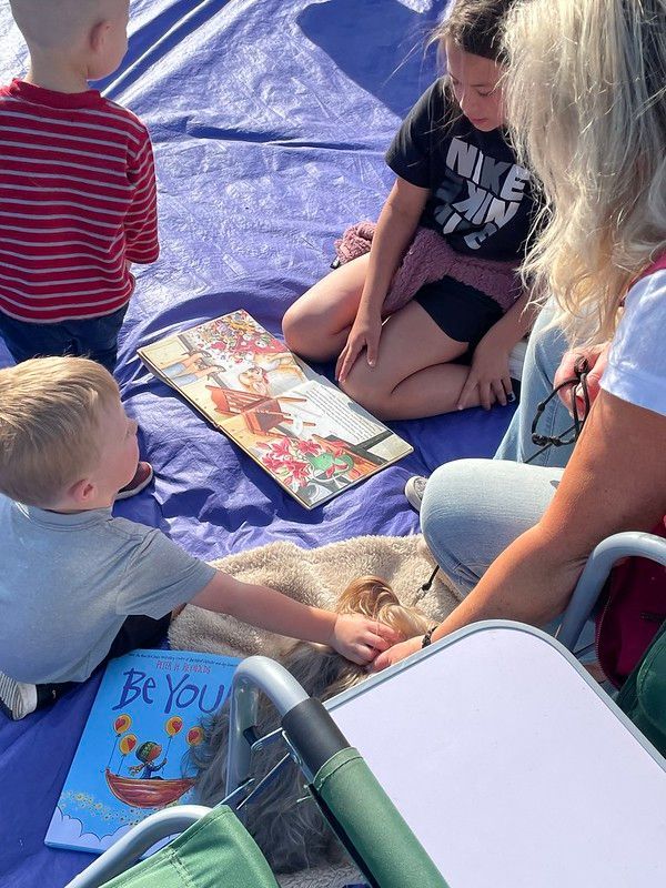 Children and an adult sit on a blue tarp, reading books outdoors.