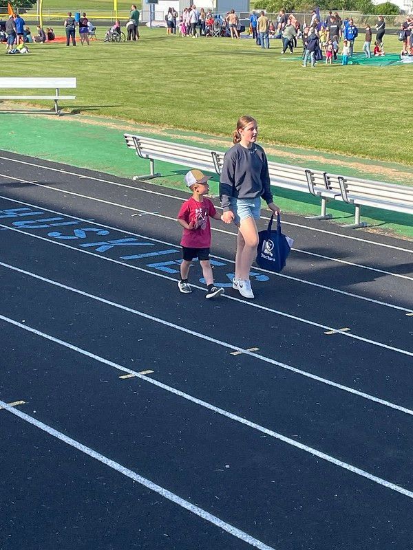 A person and a child walk on a track, holding hands. Other people are in the background on a green field.