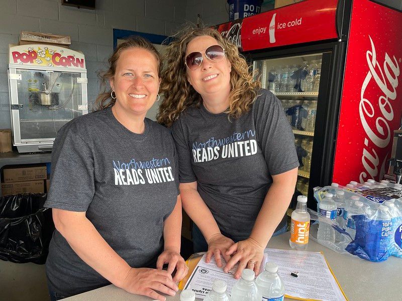 Two women wearing matching shirts, smiling, behind a concession stand, with popcorn machine and Coca-Cola cooler.