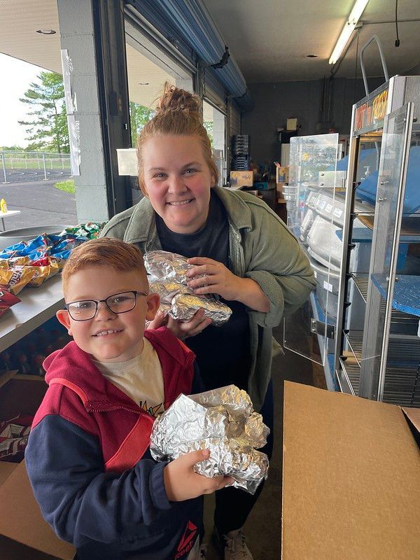 Woman and child holding foil-wrapped food, smiling, inside a room with shelves and a window.