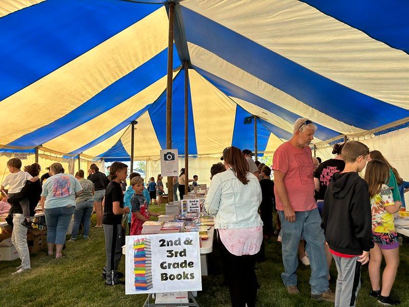 Book sale under a blue and white striped tent. People browse tables of books, outdoors.