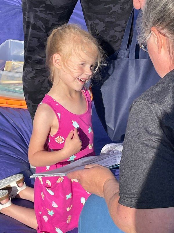 Girl in pink dress smiles, kneeling, reading book with older person, outdoors.