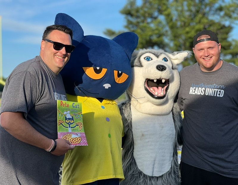 Two men pose with Pete the Cat and a wolf mascot holding a Pete the Cat book.