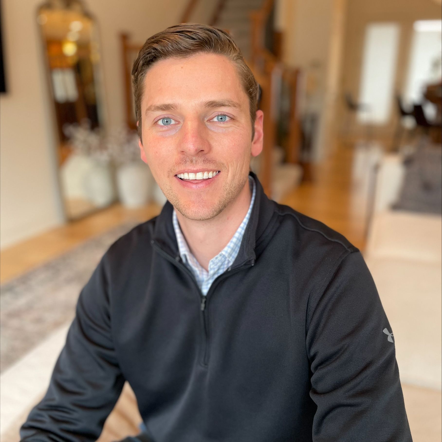 Man smiling, looking at the camera, wearing a black jacket over a patterned shirt, home interior in background.