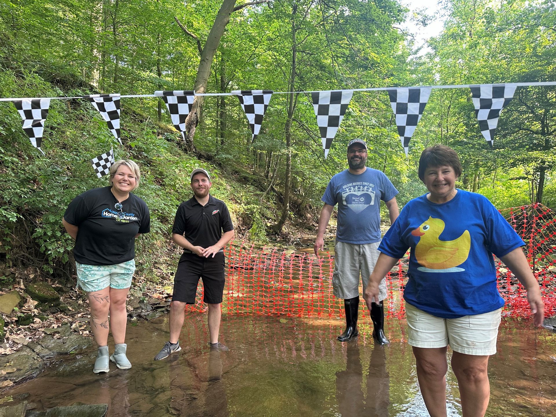 Four people standing in a stream under checkered flags, smiling. Surrounded by trees and red flowers.