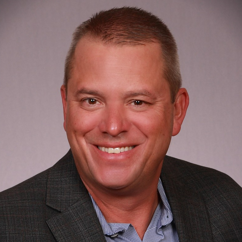 Smiling man in a blue shirt and dark blazer against a gray background.