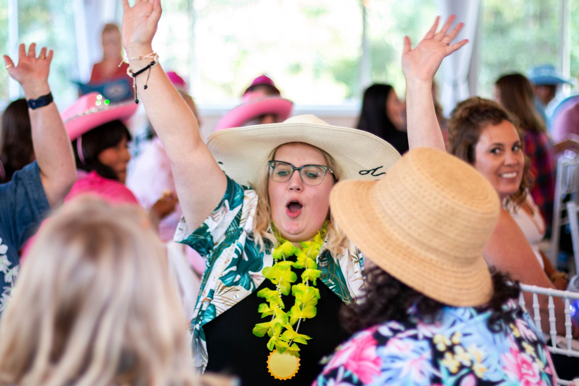 Woman with arms raised and mouth open, wearing a straw hat, lei, and floral shirt, at a party.