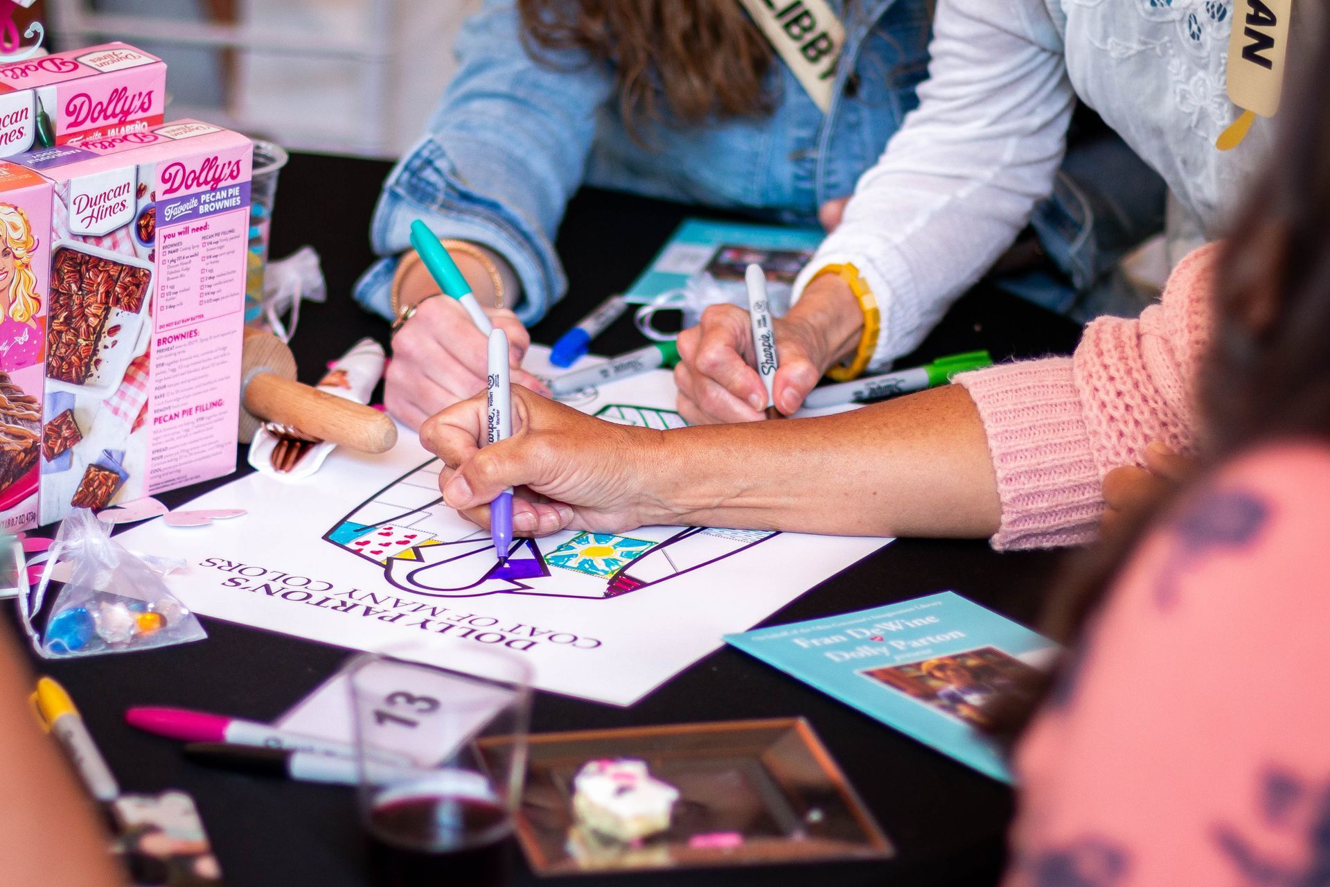People coloring at a table; markers and coloring page visible, along with a snack.
