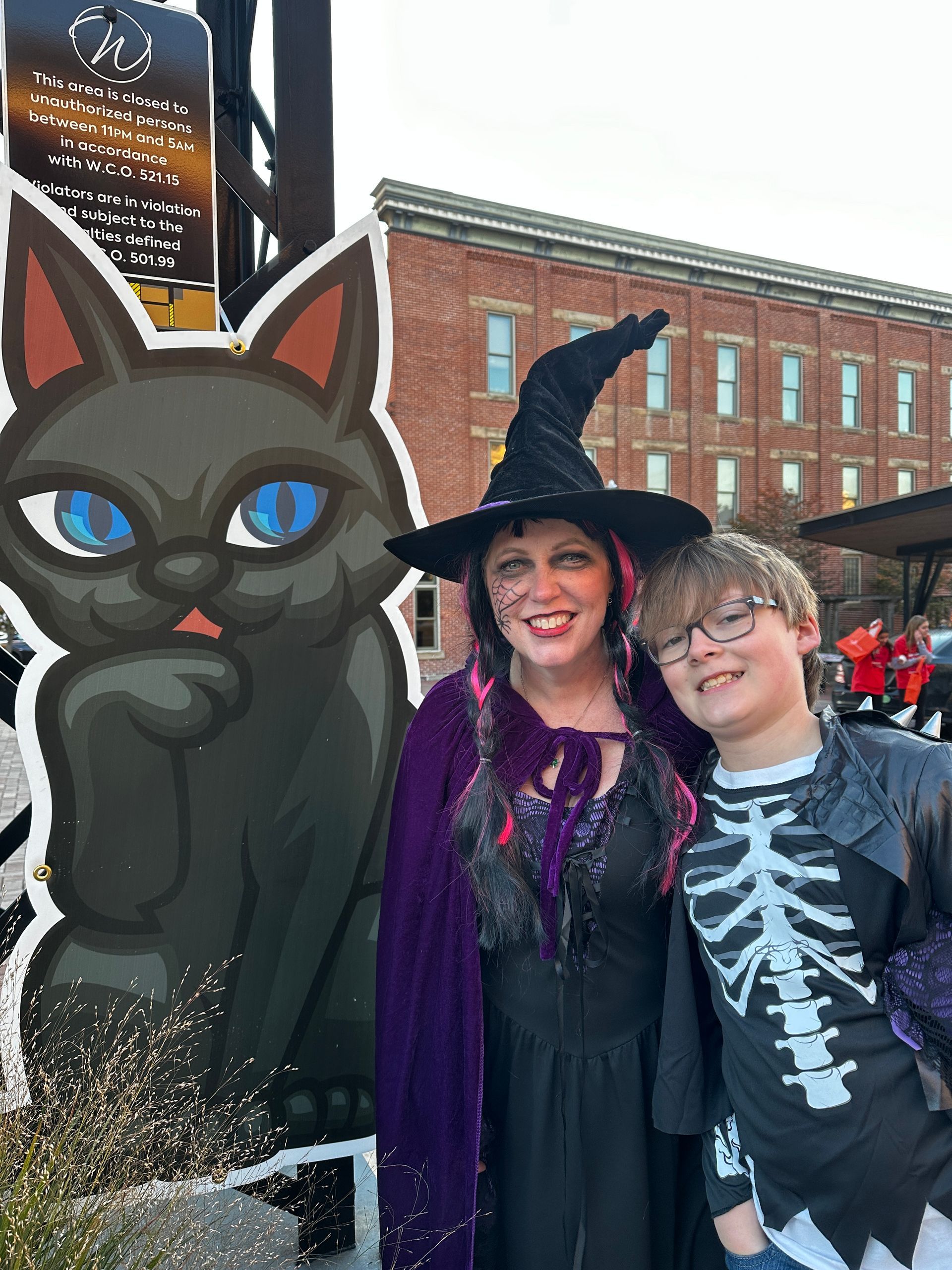 Woman and child in Halloween costumes pose next to a black cat cutout.