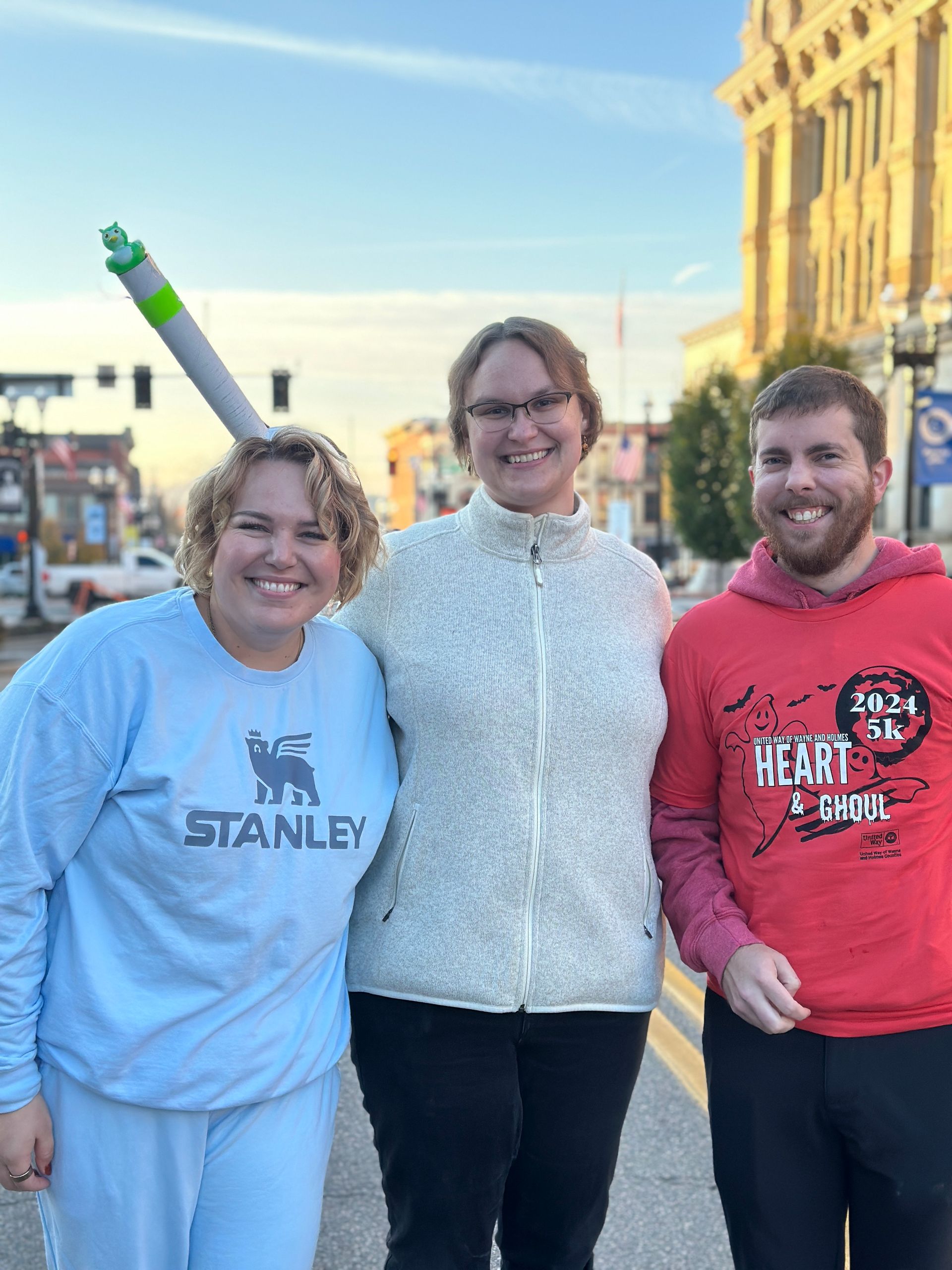 Three people posing on a street; one wears a Stanley sweatshirt and a prop, others smile.