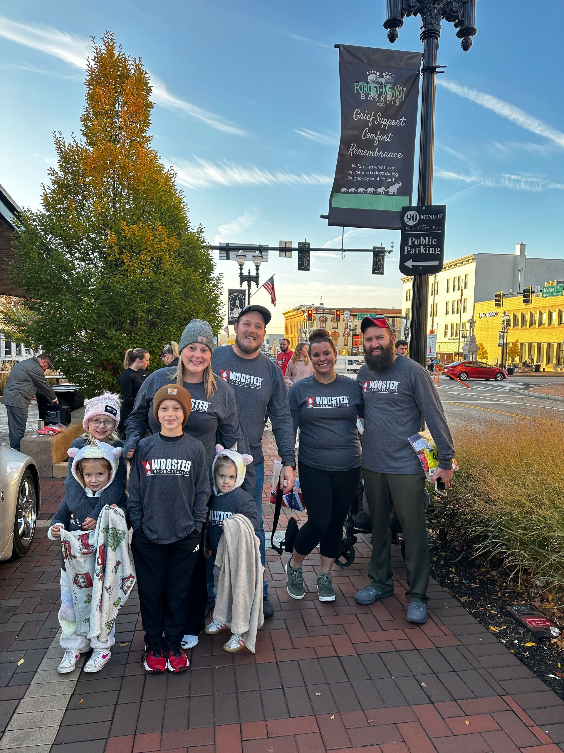 Group of people in matching shirts with kids in costumes, posing on a sidewalk near a street with fall foliage.
