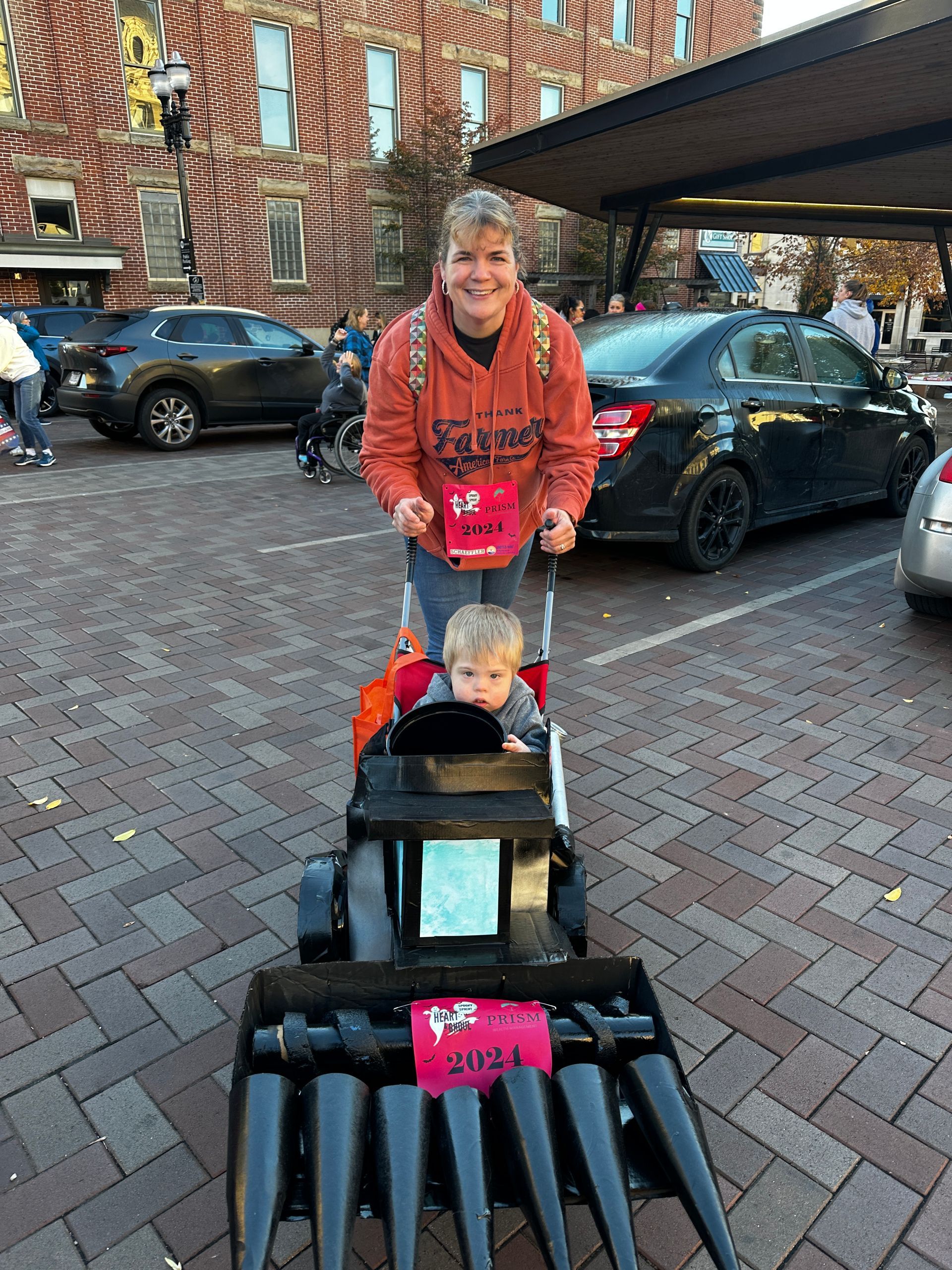 Woman pushes child in a lawnmower-themed stroller in a parking lot. Buildings in the background.