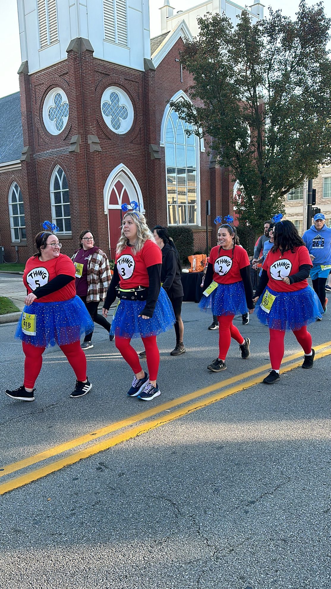 People in red shirts, blue tutus, and hats walk down a street, past a brick building.