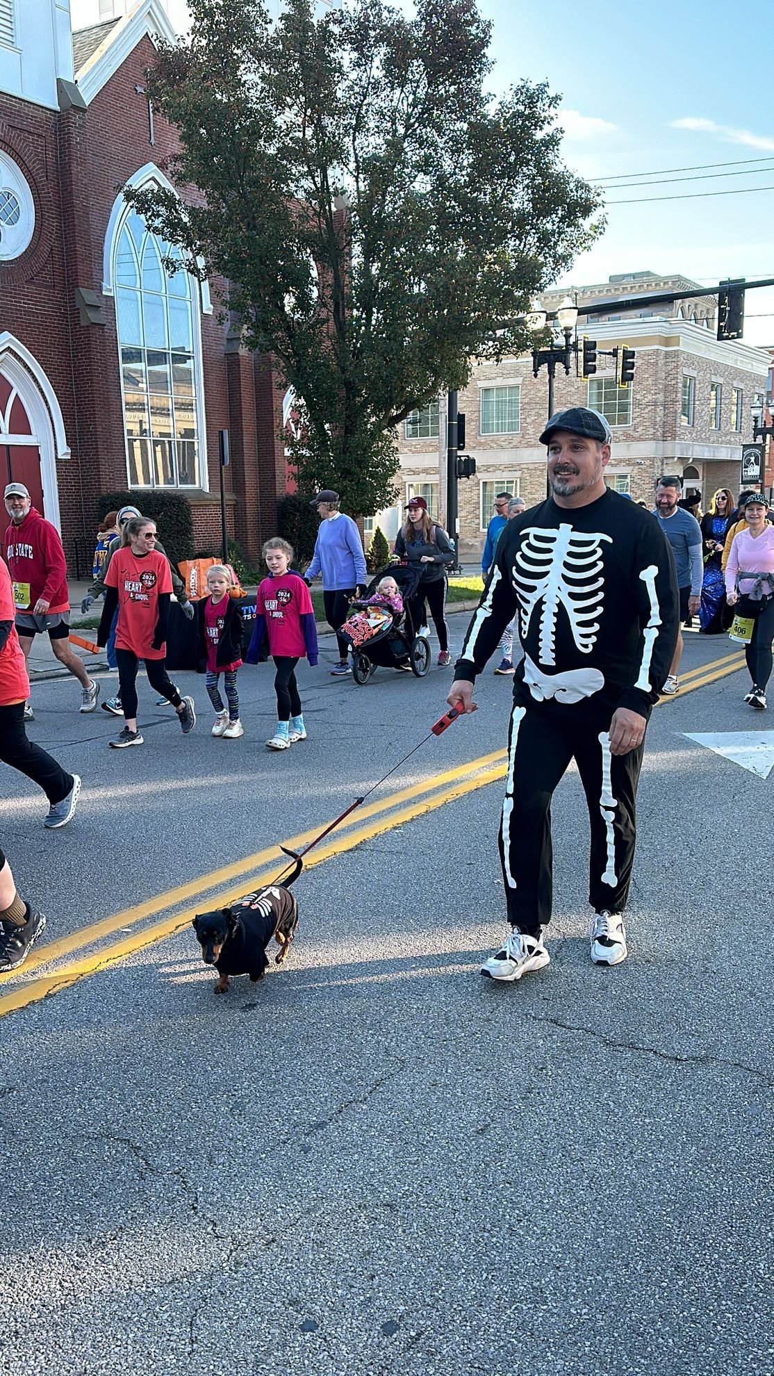 Man in skeleton costume walks a small dog in a street parade.