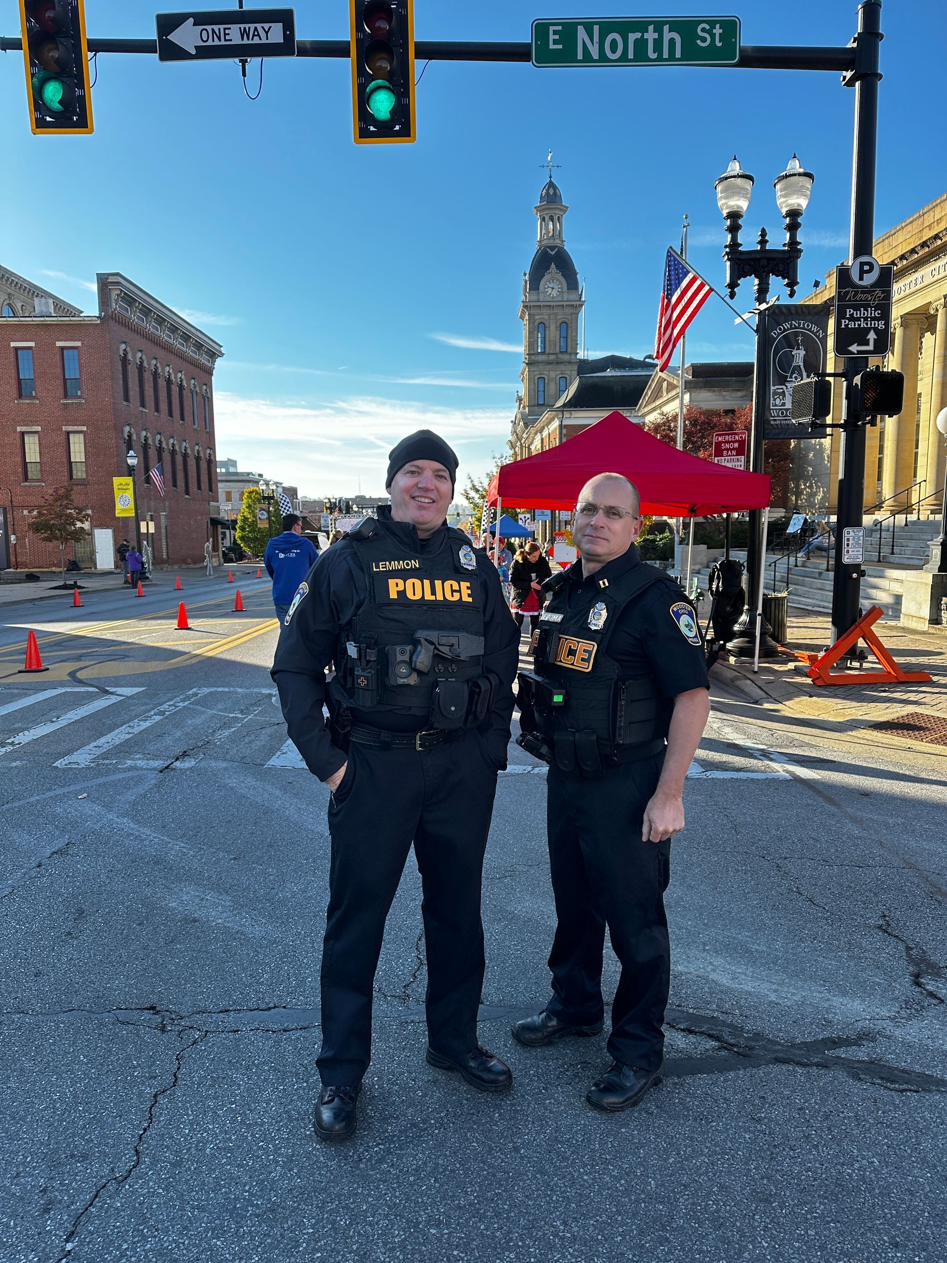Two police officers stand in a street, one smiling, in front of a building with a clock tower and American flags.