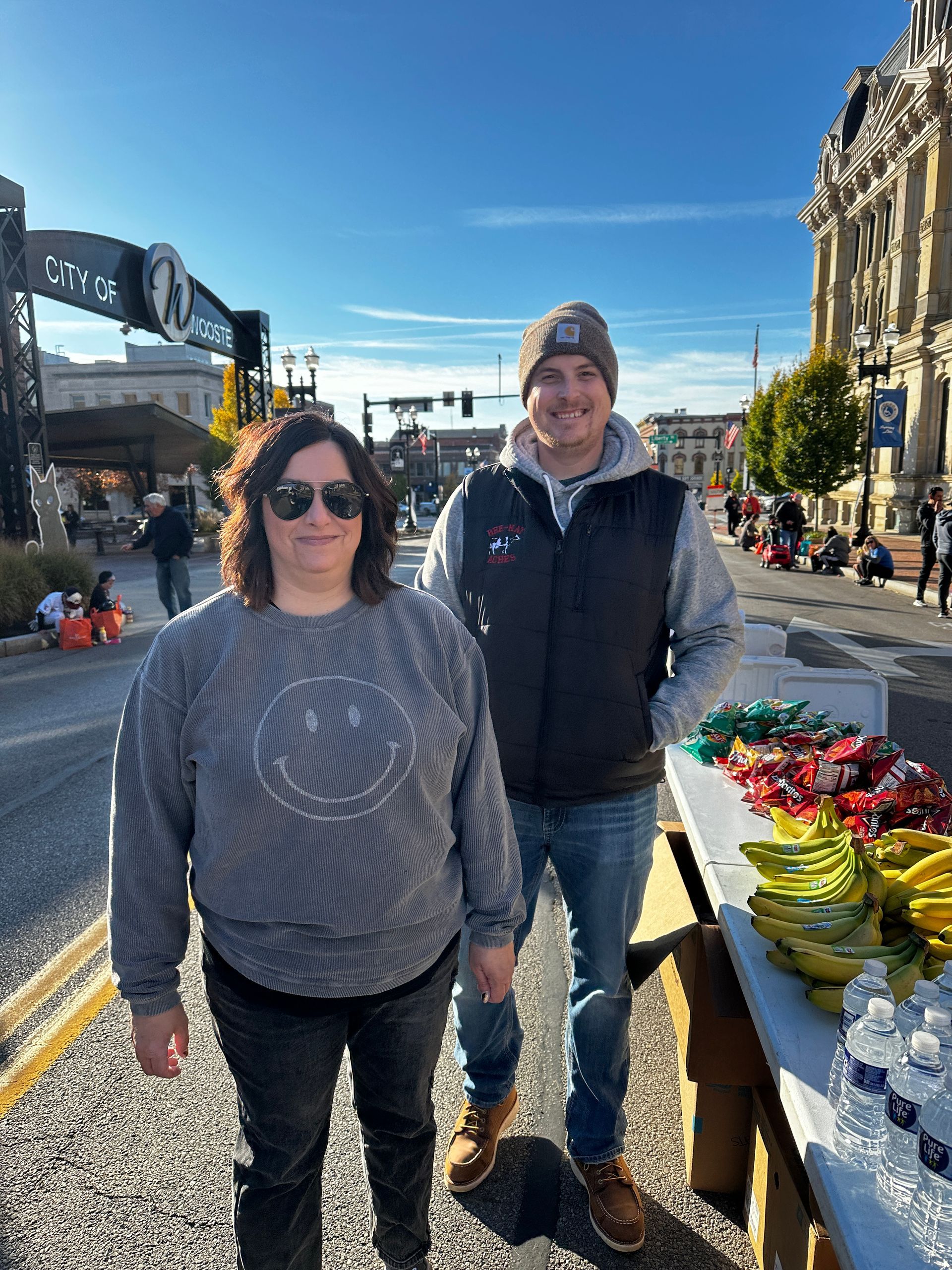 Two people stand near a table of snacks on a city street. The sky is blue.