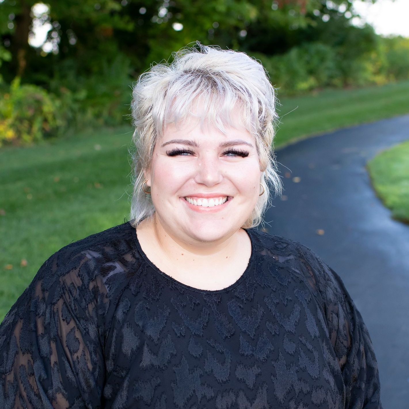 Woman with blonde hair smiles, wearing a black lace shirt, in front of a curved path and green landscape.