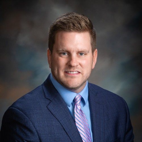 Man in blue suit and light purple tie smiles. Dark background.