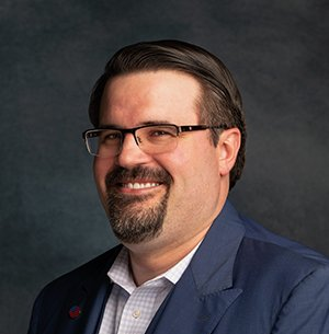 Man with glasses, beard, and blue blazer smiles, looking at the viewer.