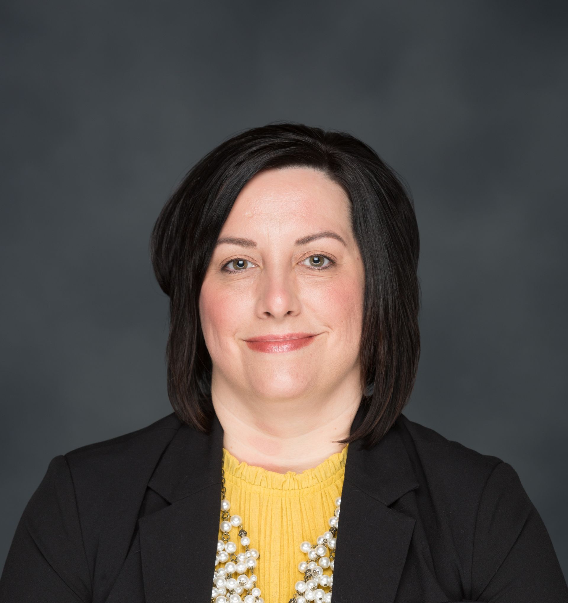 Woman with dark hair in black blazer, yellow shirt, and pearl necklace. Smiling, gray backdrop.